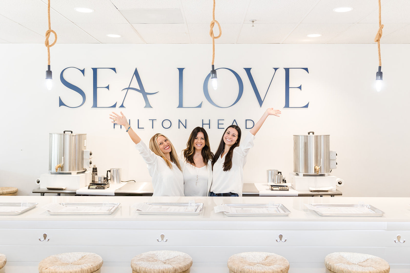 Three cheerful individuals standing behind an ice cream counter at "sea love hilton head," with arms raised in a welcoming gesture.