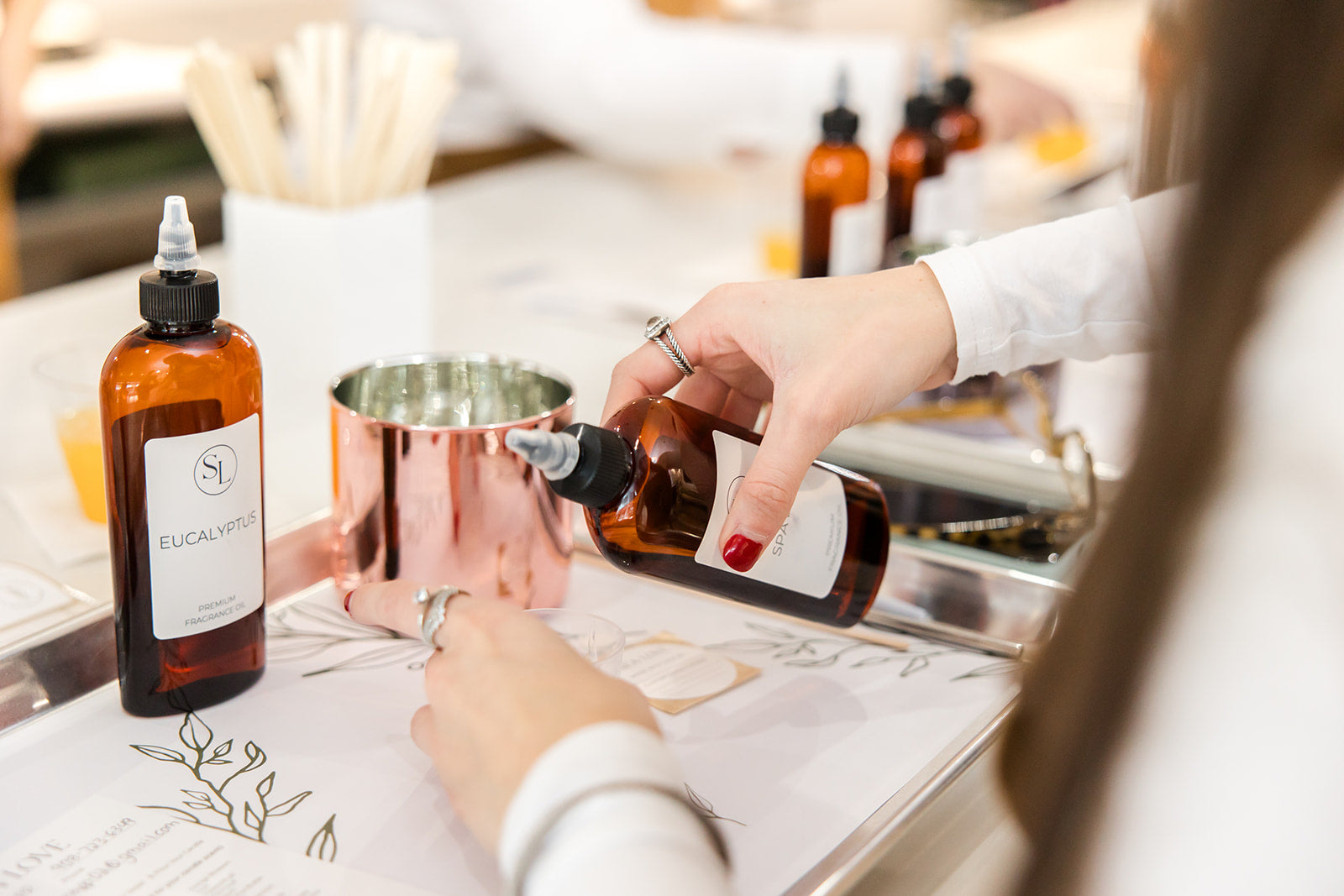 A person carefully dispensing a liquid from a brown glass bottle into a metallic measuring cup at a well-organized workstation with various bottles and ingredients on display, hinting at a process of precise formulation or bespoke product creation.