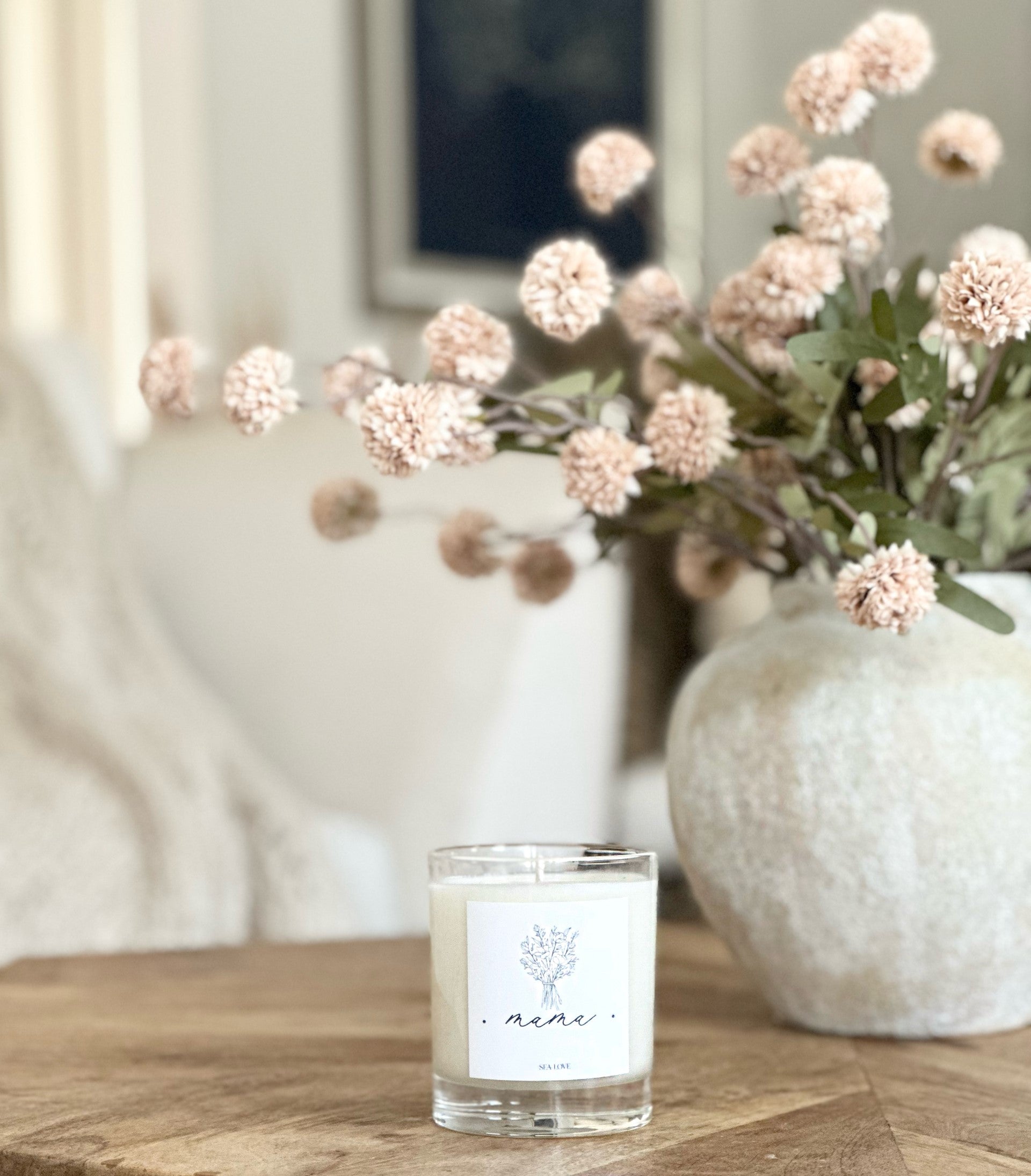 A scented candle with a simple label "mama" on a wooden table, framed by a vase of delicate pink flowers in the softly blurred background, creating a cozy atmosphere.