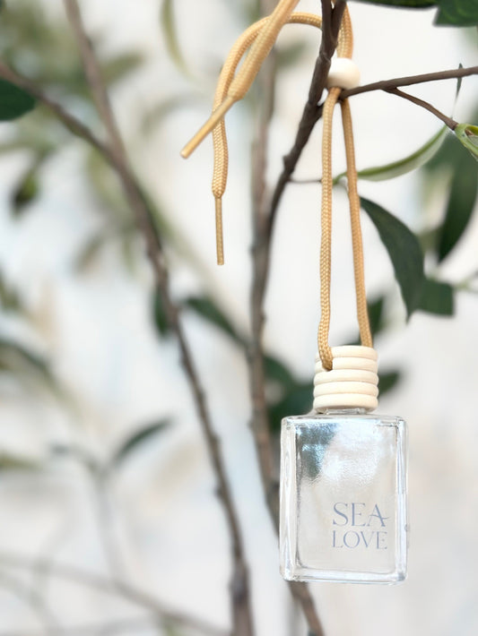 A translucent perfume bottle with the label "sea love" hanging from a beige ribbon on a green leafy branch, with a blurred background suggesting a serene and natural setting.
