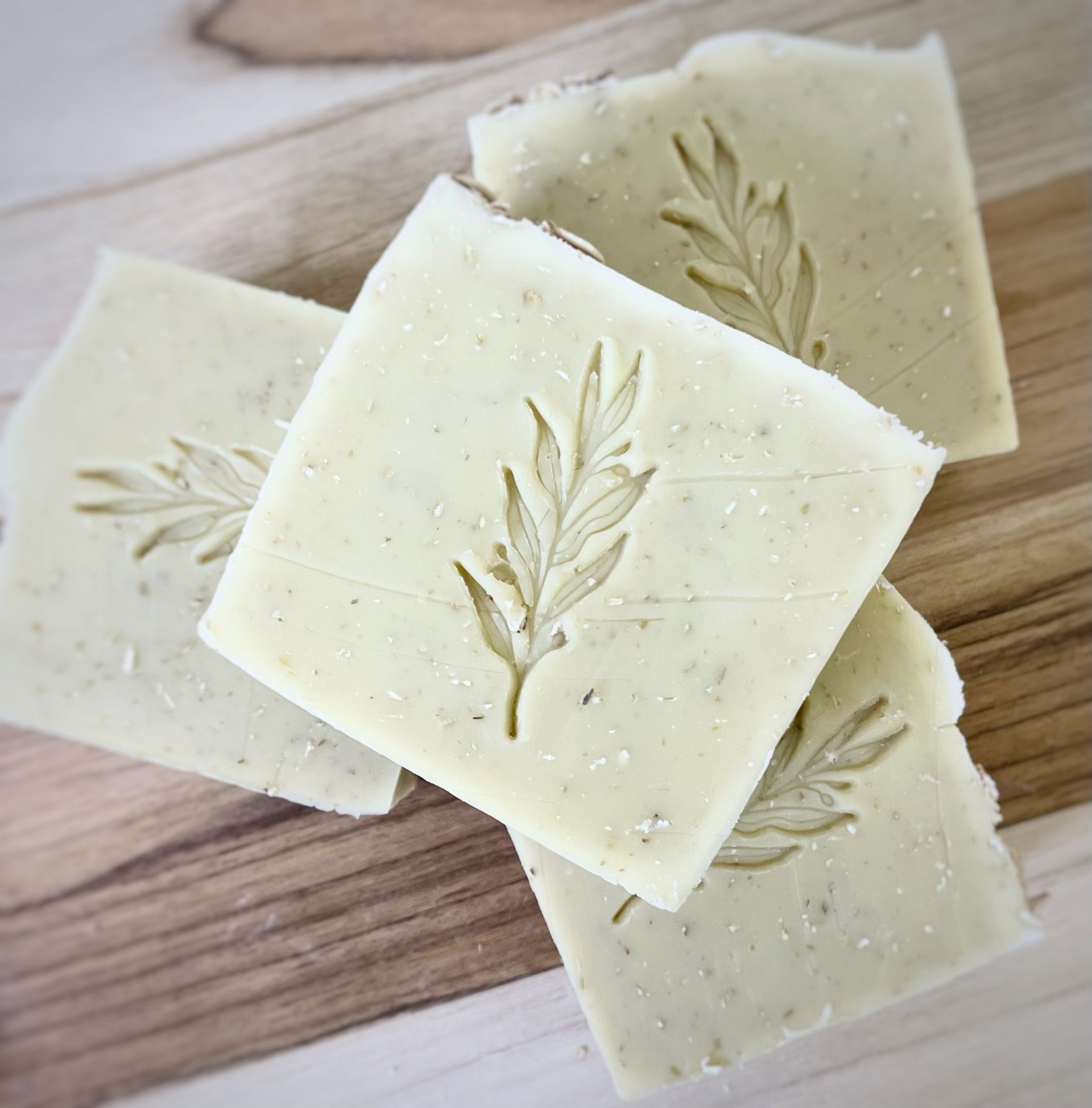 Four square artisanal soaps with an embossed leaf design, arranged in a layered pattern on a wooden surface.